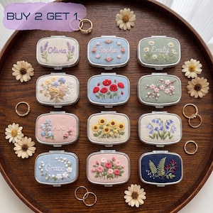 Puede incluir: Doce pastilleros rectangulares de metal con tapas de tela bordada que presentan diseños florales y nombres como Olivia, Sophie, Emily, Ava y Grace. Los estuches están dispuestos sobre una bandeja de madera con flores blancas y anillos dorados.