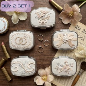 Puede incluir: Una colección de espejos compactos rectangulares con marco plateado y diseños bordados de flores y anillos. Los espejos están dispuestos sobre una superficie de madera con barras de labios, anillos y flores. El texto "BUY 2 GET 1" es visible.