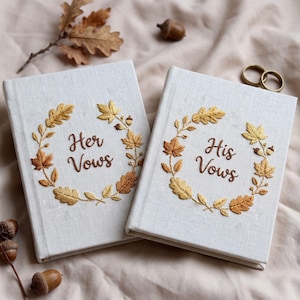 Puede incluir: Dos libros cubiertos de tela color hueso con coronas de hojas de otoño bordadas en oro y marrón. Un libro presenta el texto bordado en marrón "Her Vows" y el otro "His Vows" en el centro. Los libros están rodeados de bellotas y hojas de roble.