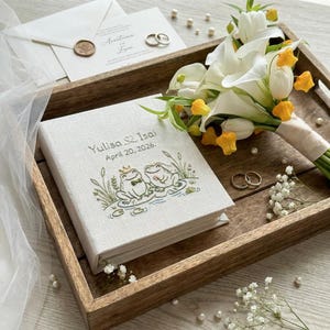 以下が含まれることがあります： 刺繍されたカエル、白いカラーと黄色の花のブーケ、結婚指輪、招待状が描かれた、リネンのフォトアルバムを特徴とする結婚をテーマにした静物画。アルバムには名前と日付が記載されています。