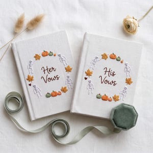 May include: Two off-white fabric-covered books with embroidered wreaths of orange pumpkins, green pumpkins, autumn leaves, and white skeletons. One book reads Her Vows and the other reads His Vows in brown script. A sage green ribbon and ring box.