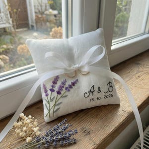 May include: A white linen ring pillow with embroidered lavender sprigs and the initials "A & B" with the date "15.10.2025" in black script. Two gold rings are attached with a white ribbon. The pillow is on a wooden surface.