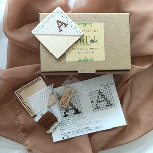 Può includere: Un kit per ricamo a punto croce con la lettera "A" ricamata su tessuto bianco. Il kit include una scatola di cartone, filo, un ago e istruzioni. Il kit è fatto a mano e include il testo "Yuliya Duinova YuinkHandMade".