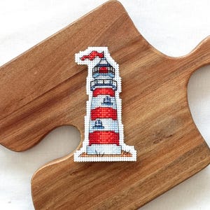 May include: A white cross-stitch pattern of a red and white striped lighthouse with a red flag blowing in the wind. The lighthouse has three windows and is sitting on a patch of sand.