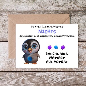 Könnte beinhalten: Eine Grußkarte mit einer Cartoon-Pinguin-Illustration. Der Text auf Deutsch lautet: „Du hast dir mal wieder nichts gewünscht, also musste ich kreativ werden.“ Auf einem Holzgrund.
