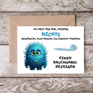 Könnte beinhalten: Grußkarte mit einer blauen Monsterillustration und dem Text "DU HAST DIR MAL WIEDER NICHTS GEWÜNSCHT, ALSO MUSSTE ICH KREATIV WERDEN. EINEN BAUCHNABEL REINIGER" auf Holzgrund.