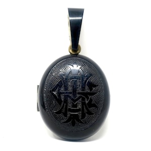 May include: A black oval pendant with an engraved monogram design. The pendant has a loop at the top for a chain and a small clasp on the side. The pendant is made of a dark material and has a polished finish.
