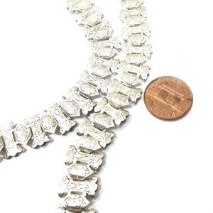 May include: Two silver chains with a decorative, floral pattern. The chains are laid out in a zig-zag pattern and are about the size of a US penny.