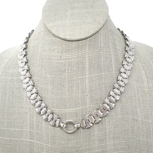 May include: A silver chain necklace with a textured, patterned design. The chain is made of interlocking links and has a simple, round clasp.
