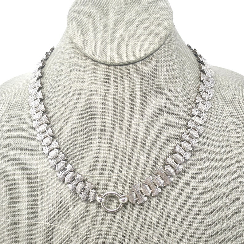 May include: A silver chain necklace with a textured, patterned design. The chain is made of interlocking links and has a simple, round clasp.