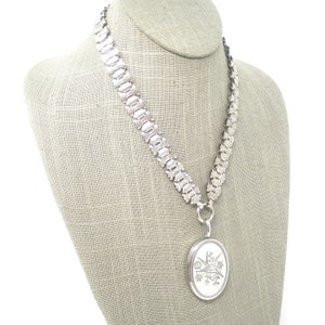 May include: A silver chain necklace with a silver locket pendant. The locket has an engraved design of a bird and flowers.