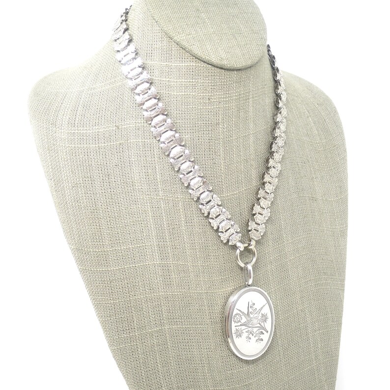 May include: A silver chain necklace with a silver locket pendant. The locket has an engraved design of a bird and flowers.