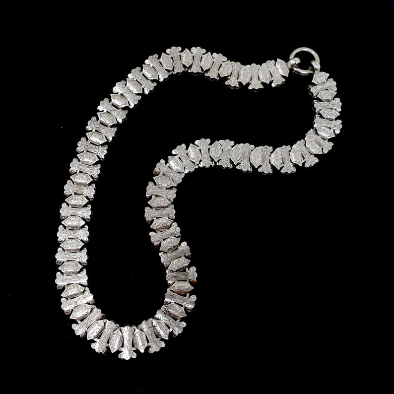 May include: A silver chain necklace with a geometric pattern. The chain is made up of small, interlocking links that form a repeating design.