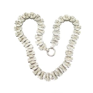 May include: A silver chain necklace with a delicate, intricate design. The chain is made up of small, linked oval shapes with a textured surface.