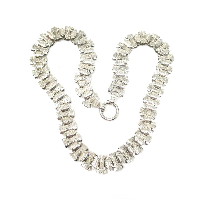 May include: A silver chain necklace with a delicate, intricate design. The chain is made up of small, linked oval shapes with a textured surface.