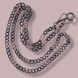 Könnte beinhalten: Eine silberfarbene Taschenuhrkette mit Karabinerverschluss und Ring. Die Kette hat eine Mischung aus Curb- und ovalen Gliedern in einem zweifarbigen Design. Die Kette wird auf einem hellrosa Hintergrund präsentiert.