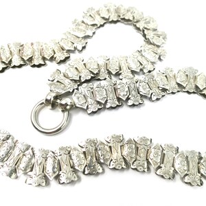 May include: A silver chain necklace with a detailed floral pattern. The chain is made of interlocking links and has a clasp.