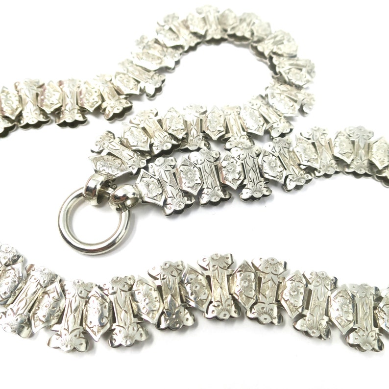 May include: A silver chain necklace with a detailed floral pattern. The chain is made of interlocking links and has a clasp.