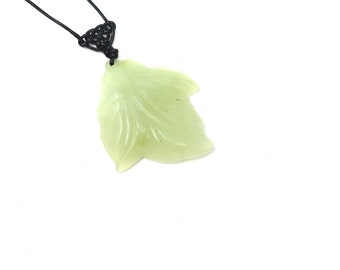 May include: A light green leaf-shaped pendant necklace. The pendant is carved with leaf details and hangs from a black cord. The cord is knotted at the top. The background is white.
