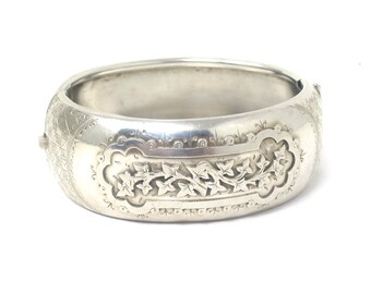 Puede incluir: Un brazalete de plata con un diseño floral intrincado. El brazalete está grabado con un patrón repetitivo de hojas y vides.