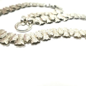 May include: A silver chain necklace with a decorative, ornate design. The chain is made of interlocking links that are shaped like small, elongated flowers. The necklace has a clasp that is a simple ring.