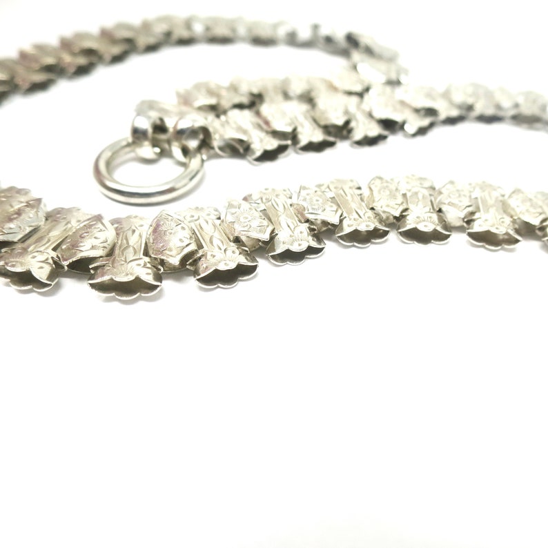 May include: A silver chain necklace with a decorative, ornate design. The chain is made of interlocking links that are shaped like small, elongated flowers. The necklace has a clasp that is a simple ring.