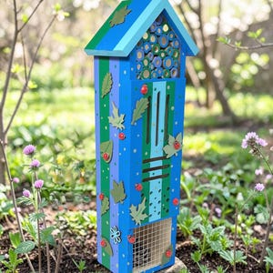 May include: A tall, rectangular wooden insect hotel painted in shades of blue and green with vertical stripes. The structure features a gabled roof, decorative green leaf cutouts, small red ladybug accents, and a wire mesh compartment at the base.