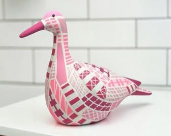 May include: A decorative bird figurine featuring a geometric pattern in various shades of pink, white, and magenta. The bird has a smooth, rounded body, a long neck, and a solid pink beak, with a surface covered in intersecting lines and shapes.