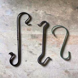 May include: Three metal hooks in varying colors: black, dark gray, and green. Each hook has a curved top and bottom, designed for hanging items. The hooks are displayed on a wooden surface.