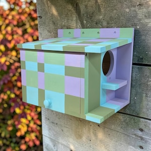 Könnte beinhalten: Ein hölzernes Vogelhaus mit einem geometrischen Schachbrettmuster in den Farben Hellblau, Lavendel und Olivgrün. Die Struktur umfasst ein schräges Dach, ein kreisförmiges Eingangsloch, eine kleine Landestange und einen runden Knopf an der Vorderseite.