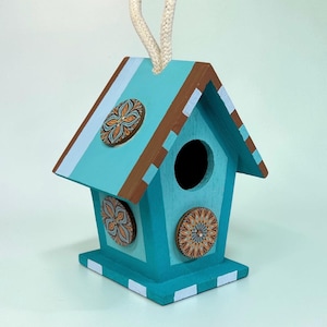 Könnte beinhalten: Ein türkisfarbenes Vogelhaus aus Holz mit braunem Dach und dekorativen floralen Akzenten. Das Vogelhaus hat ein rundes Eingangsloch und ein weißes Seil zum Aufhängen. Das Design umfasst Streifen und kreisförmige Verzierungen in Orange- und Brauntönen.