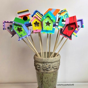 May include: A bouquet of colorful wooden birdhouses with painted details, each with a different design and color scheme. The birdhouses are attached to wooden sticks and arranged in a vase.