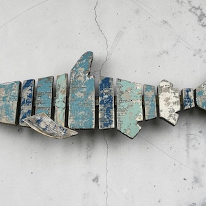 Puede incluir: Escultura de tiburón de madera con aspecto envejecido y desgastado. El tiburón está construido con piezas rectangulares de madera pintadas en tonos de azul, blanco y gris. La escultura está montada en una pared gris claro.