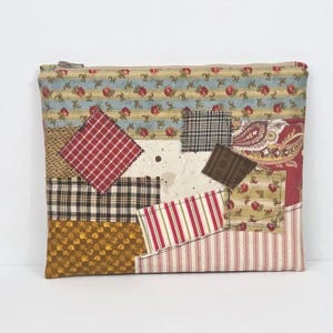 May include: A patchwork zippered pouch featuring a variety of fabric patterns. The design includes red plaid, floral, striped, and paisley fabrics in shades of red, blue, brown, and gold. The pouch has a beige zipper and a rectangular shape.
