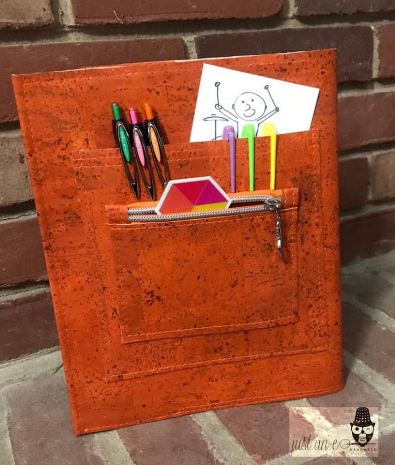 May include: Orange cork notebook with a zippered pocket and a small pocket for pens and pencils. The notebook has a colorful geometric sticker on the front.