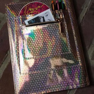 May include: A holographic pink and silver folder with a zippered pocket and a hexagonal pattern. The folder has a CD and a card inside the pocket. There are also three pens in the pocket.