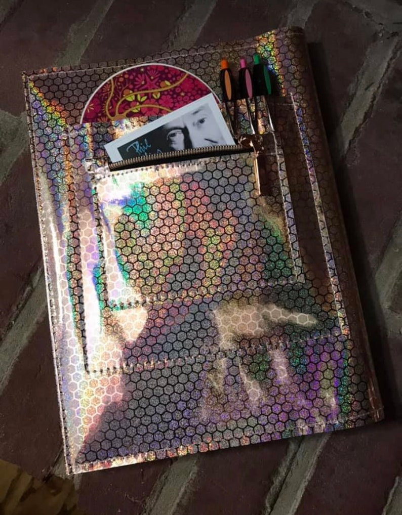 May include: A holographic pink and silver folder with a zippered pocket and a hexagonal pattern. The folder has a CD and a card inside the pocket. There are also three pens in the pocket.