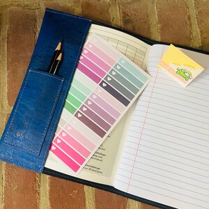 May include: A blue cork notebook with a pencil holder and a sheet of colorful heart stickers. The notebook has lined pages and a sticker that says "It's well be cool".