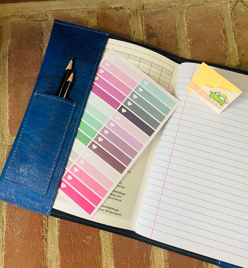 May include: A blue cork notebook with a pencil holder and a sheet of colorful heart stickers. The notebook has lined pages and a sticker that says "It's well be cool".