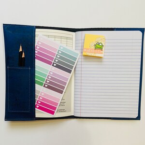 May include: A blue notebook with a corkboard cover and a zippered pocket. The notebook has lined pages and a sticker with a frog on it. The notebook also has a sheet of colorful stickers with hearts.