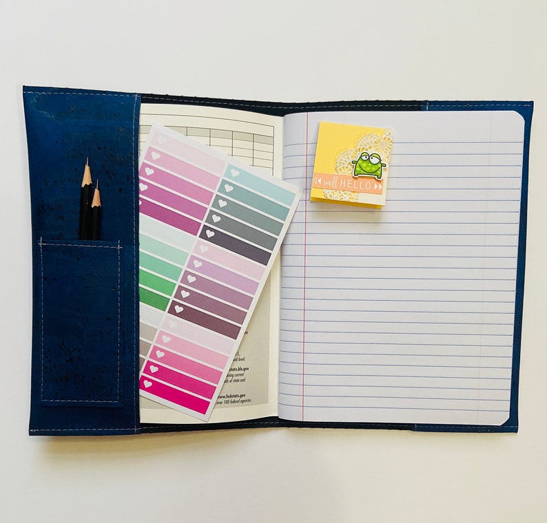 May include: A blue notebook with a corkboard cover and a zippered pocket. The notebook has lined pages and a sticker with a frog on it. The notebook also has a sheet of colorful stickers with hearts.