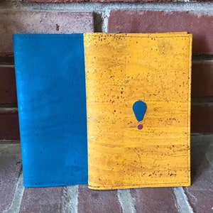 May include: A cork notebook with a blue cover on the left and a yellow cork cover on the right. The yellow cover has a blue and red accent.