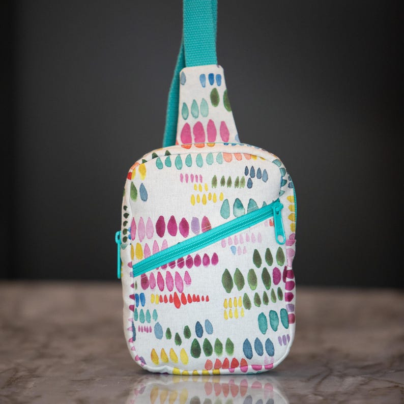 May include: A crossbody sling bag featuring a white fabric base with a colorful pattern of small, painted-style teardrop shapes in shades of pink, yellow, blue, and green. The bag includes a bright teal zipper and a matching teal strap.