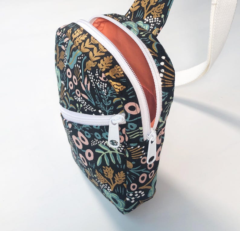 May include: A black crossbody bag featuring a floral pattern in shades of gold, pink, and teal. The bag has a white zipper closure, a white strap, and a bright orange interior lining. The exterior fabric displays a dense botanical print.