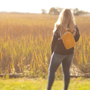 May include: A mustard-yellow sling backpack with a small front pocket is worn over the shoulder. The person wearing the backpack is standing in a field of tall, golden grass. The background is blurred, with a bright, sunny sky.
