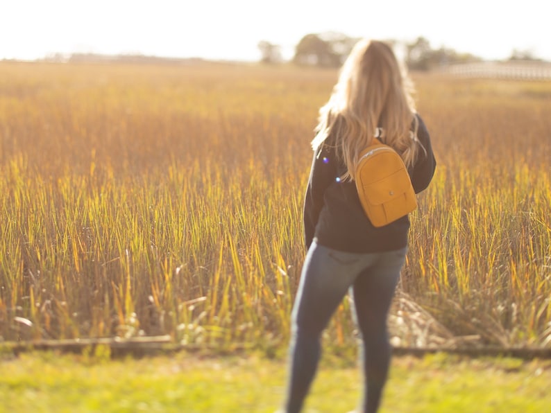May include: A mustard-yellow sling backpack with a small front pocket is worn over the shoulder. The person wearing the backpack is standing in a field of tall, golden grass. The background is blurred, with a bright, sunny sky.