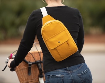 May include: A mustard yellow crossbody sling bag featuring a white strap, a main zippered compartment, and a smaller front pocket with a snap button closure. The bag is worn over the shoulder, resting against the back of a black long-sleeved shirt.