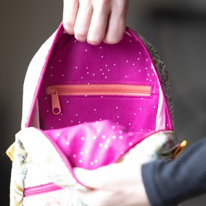 May include: A pink and white patterned backpack with a zippered pocket inside. The pocket is lined with a bright pink fabric with white polka dots.