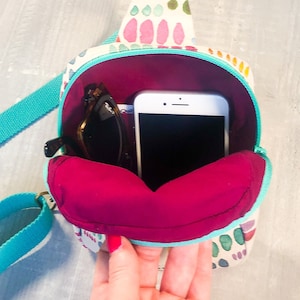 May include: A small crossbody bag with a cream-colored exterior featuring a multi-colored abstract pattern of pink, green, and orange shapes. The bag has a bright turquoise strap and zipper, with a deep magenta interior lining holding a smartphone.