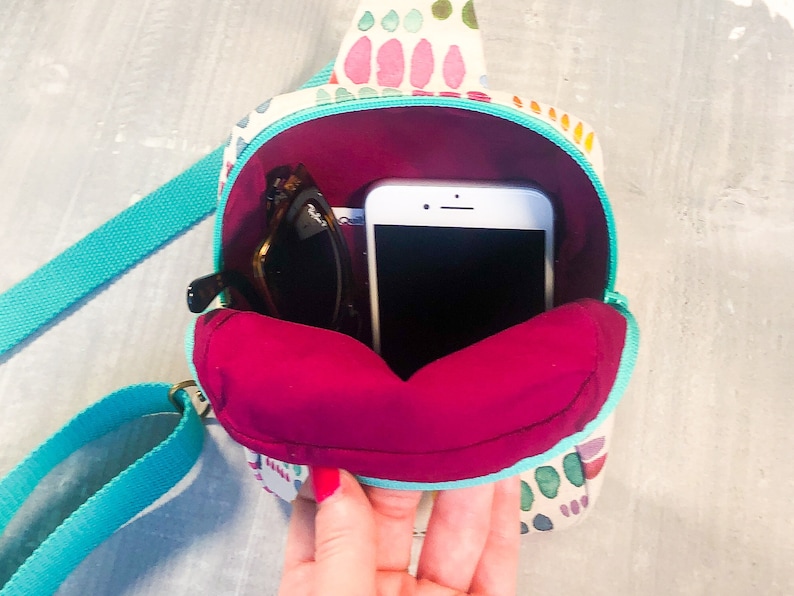 May include: A small crossbody bag with a cream-colored exterior featuring a multi-colored abstract pattern of pink, green, and orange shapes. The bag has a bright turquoise strap and zipper, with a deep magenta interior lining holding a smartphone.